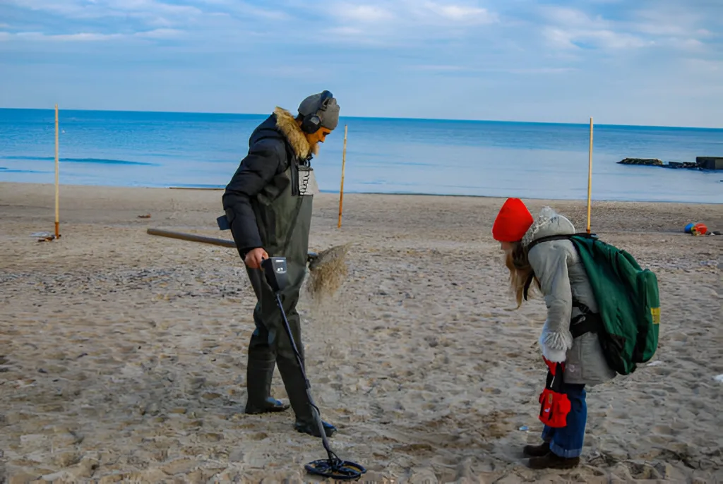 A dad and a daughter are using a metal detector to do a beach detecting.